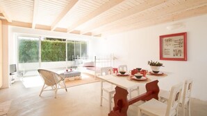 Een lichte, ruime kamer met een houten plafond, een eettafel gedekt met rood en wit servies, en een groot raam met uitzicht op groen buiten.