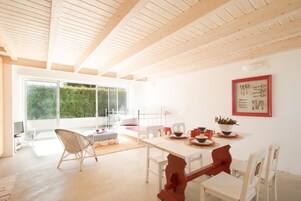 Una habitación luminosa y amplia con techo de madera, una mesa de comedor dispuesta con vajilla roja y blanca, y un ventanal que ofrece vistas a la vegetación.