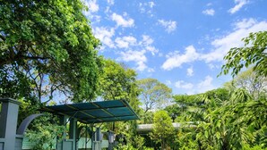 Una piscina con estructura de techo verde, rodeada de abundante vegetación y un cielo azul despejado.