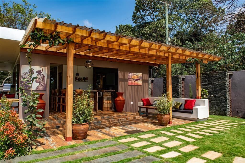 Uma área de estar coberta ao ar livre com pérgola de madeira, sofá com almofadas vermelhas e plantas em vasos.