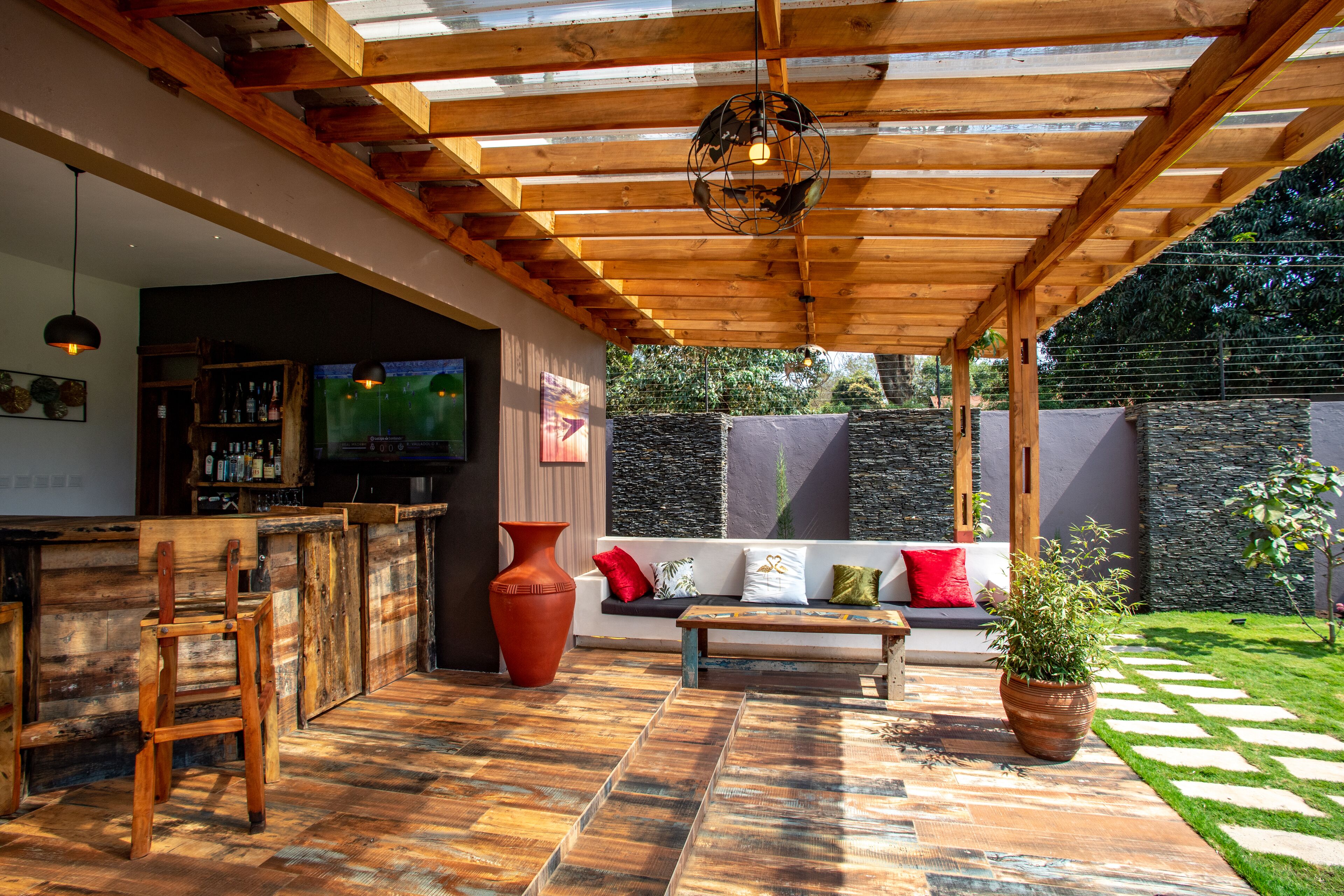 A covered outdoor area with a wooden ceiling, a bar area, a seating area with a sofa and cushions, and a potted plant.