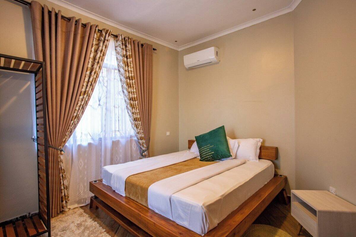A bedroom with a wooden bed, beige walls, a window with curtains, and an air conditioning unit.