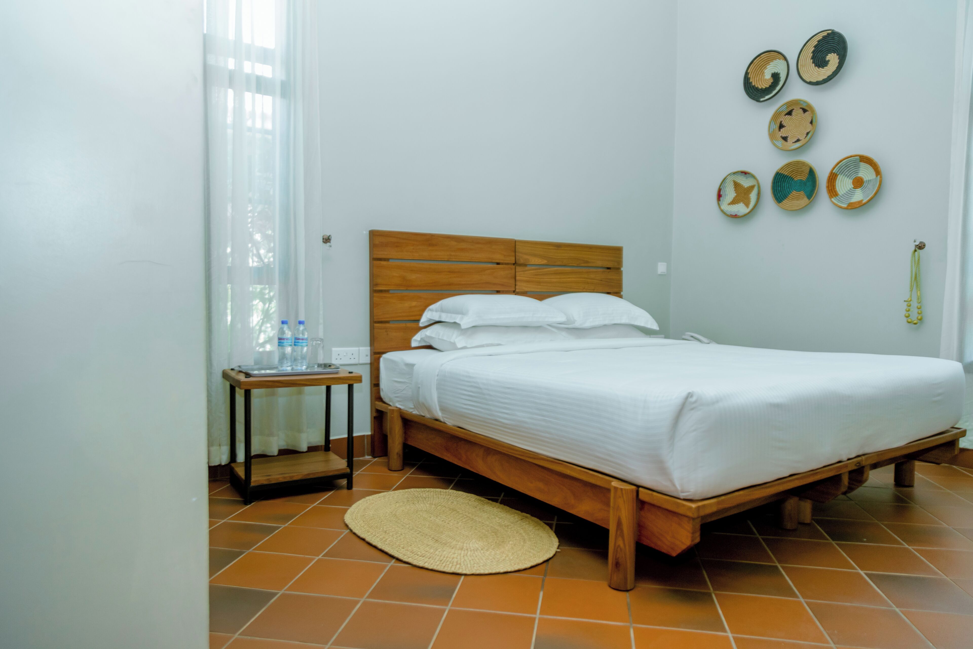 Un dormitorio con cama de madera, ropa de cama blanca, una mesita de noche pequeña, un tapete tejido y adornos en la pared.