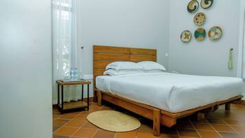Un dormitorio con cama de madera, ropa de cama blanca, una mesita de noche pequeña, un tapete tejido y adornos en la pared.
