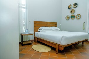 Un dormitorio con cama de madera, ropa de cama blanca, una mesita de noche pequeña, un tapete tejido y adornos en la pared.