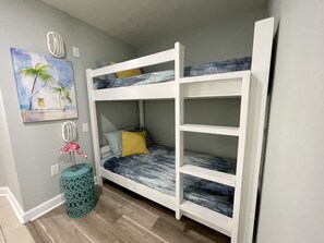 Ein Etagenbett mit blauer und weißer Matratze, einem türkisblauen Mülleimer und einem Palmen-Gemälde an der Wand.