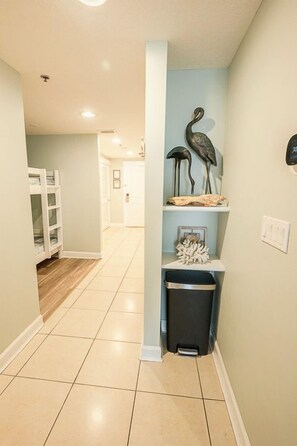 A hallway with a trash bin, a shelf with decorative items, and a door at the end.