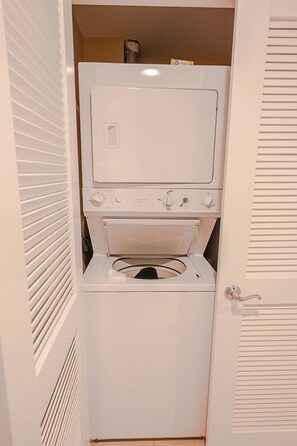 A compact laundry room with a stacked washer and dryer, a door with a handle, and a vented exhaust system. - Grand Panama Resort Condo (Panama City Beach)
