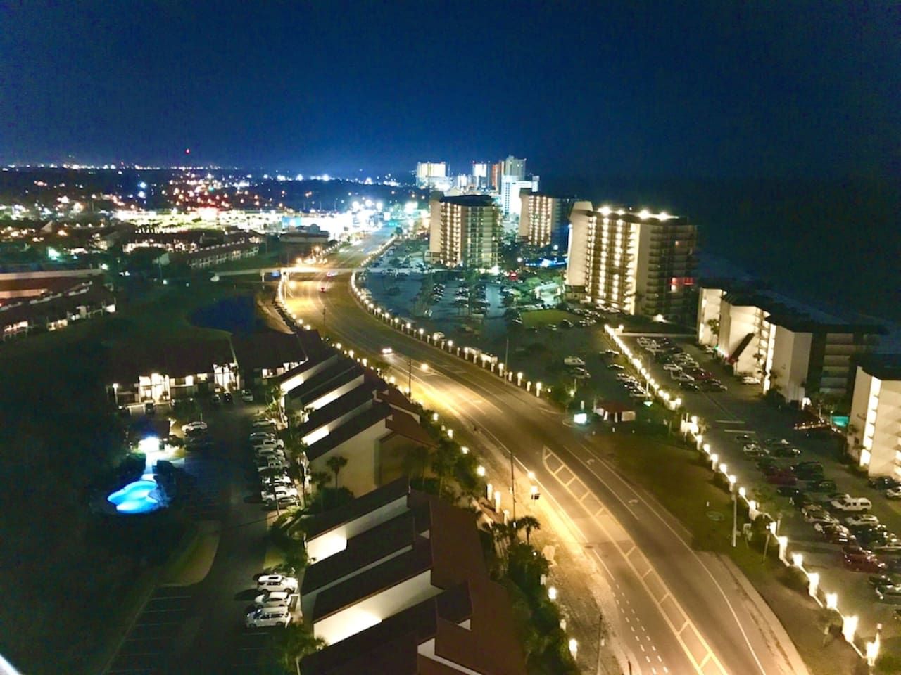A nighttime view of a coastal urban area with high-rise buildings, a curved road, and a parking lot.