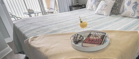 A hotel room with a bed, a bedside table with a book and a glass of drink, a chair, and a balcony view.