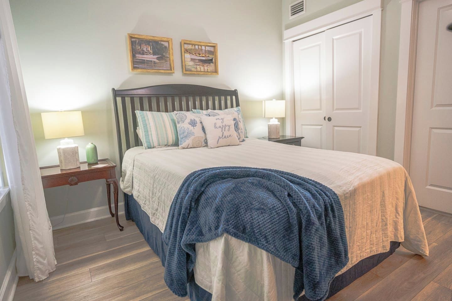 Un dormitorio con cama, mesitas de noche, una puerta y dos cuadros en la pared.