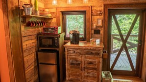 A compact kitchen with a refrigerator, a microwave, a coffee maker, and a toaster oven. There is a wooden cabinet with a window above it, and a door with a cross-brace design.