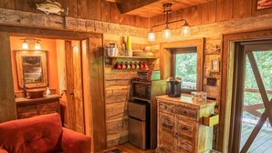 A rustic cabin interior with a kitchenette, a red sofa, wooden walls, and a view through a window.