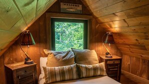 A cozy attic bedroom with a bed, two nightstands, and a window with a view of trees.