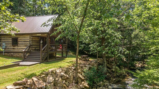 A rustic log cabin with a porch, a stone retaining wall, and a small stream.