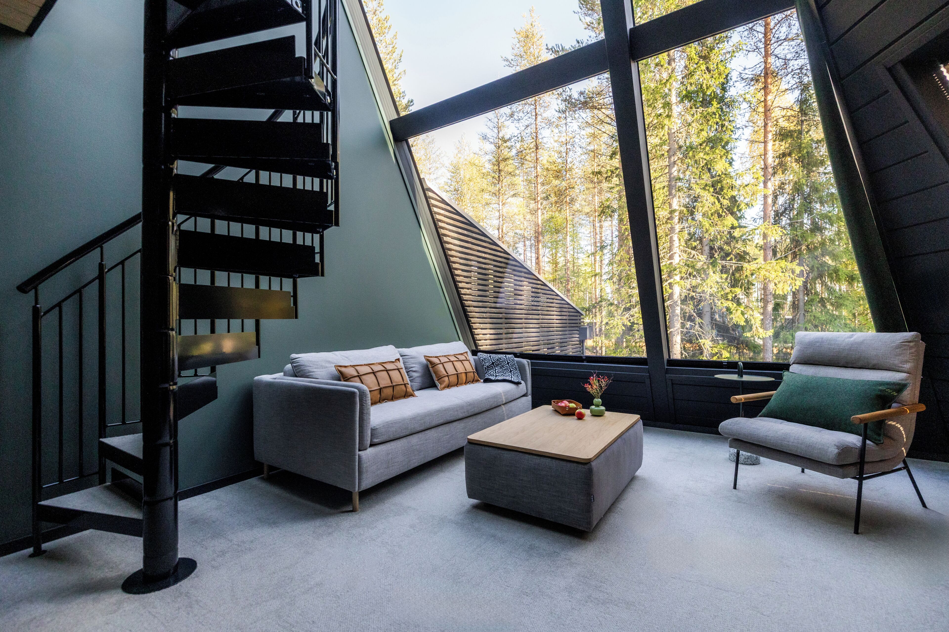 A modern interior with a grey sofa, a wooden coffee table, and a green armchair. A spiral staircase leads to an upper level.