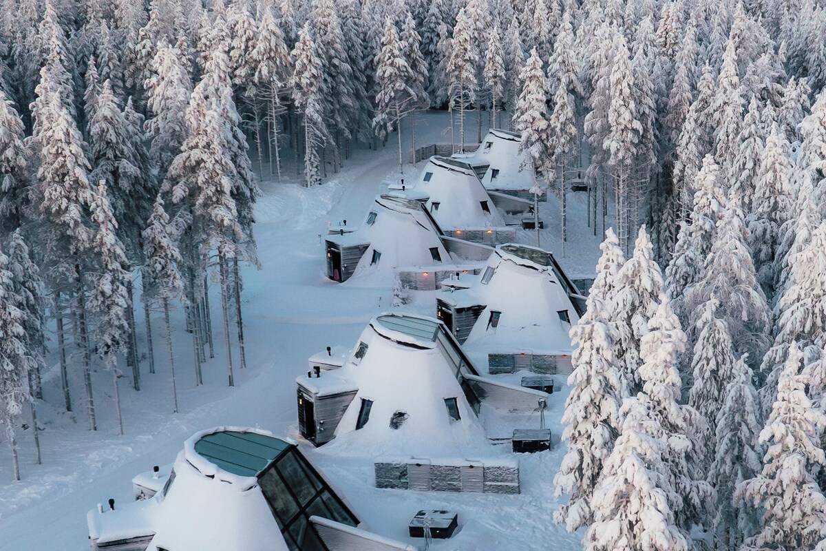 A row of snow-covered yurts in a forest, each with a sloped roof and a small porch area.