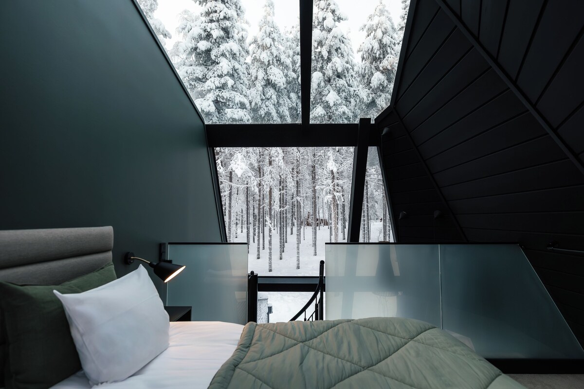 A bedroom with a bed, a nightstand, a lamp, and a view of a snowy forest through a large window.