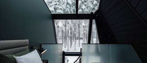 A bedroom with a bed, a nightstand, a lamp, and a view of a snowy forest through a large window.