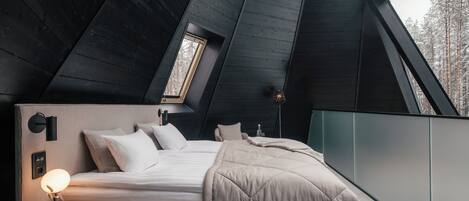 Une chambre à coucher moderne avec un plafond en pente, un lit recouvert d’une couette matelassée, une lampe de chevet et une vue sur des arbres enneigés par la fenêtre.
