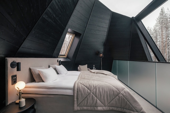 A modern bedroom with a sloped ceiling, a bed with a quilted duvet, a bedside lamp, and a view of snow-covered trees through a window.