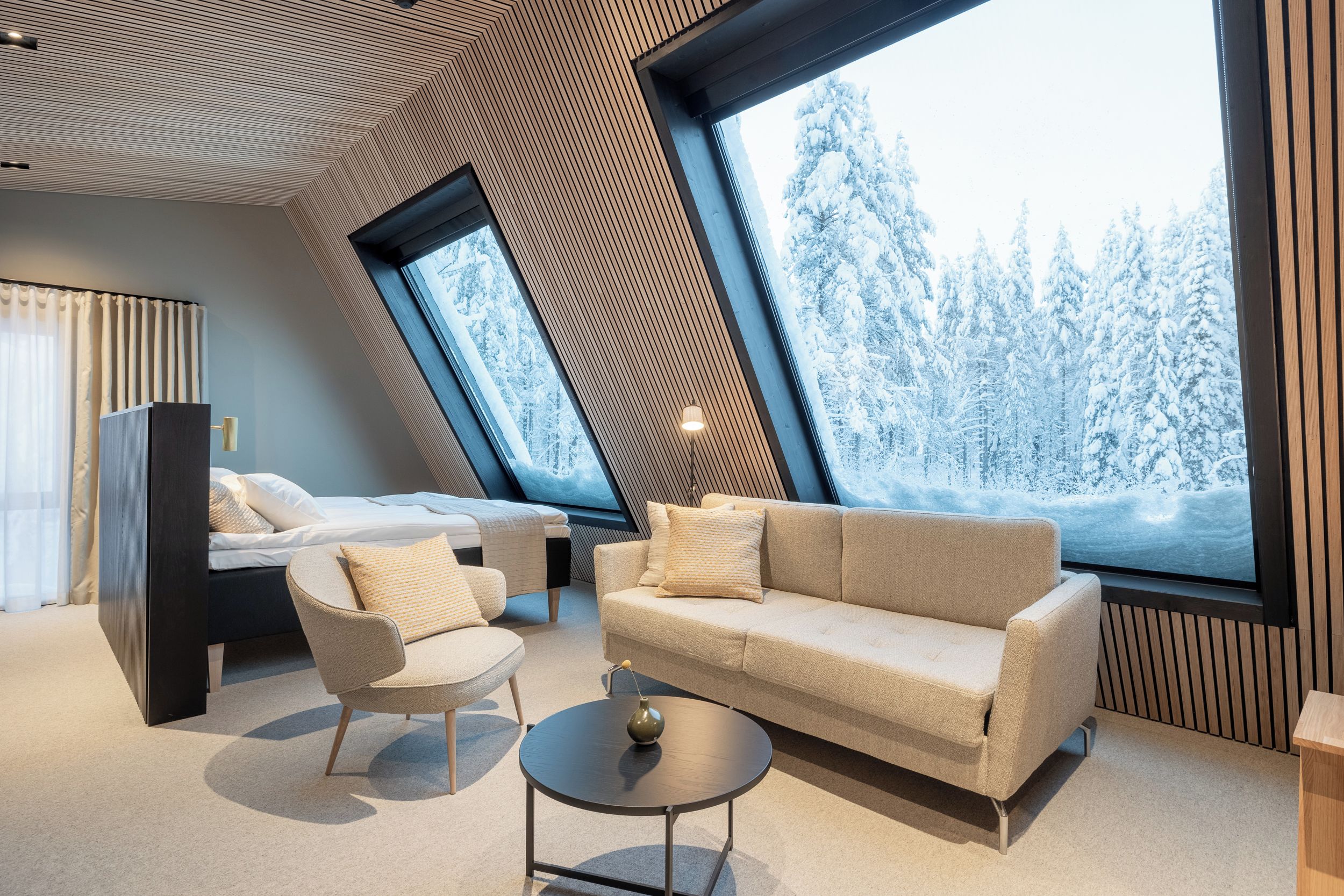 A modern interior with a sloped ceiling, a sofa, a chair, a small table, and a large window offering a snowy forest view.