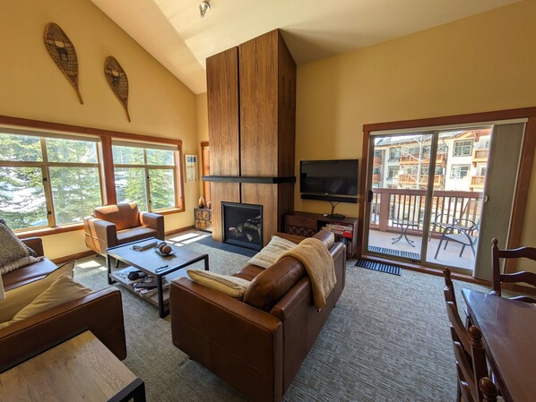A spacious living room with a fireplace, a television, and large windows offering a view of the outdoors.