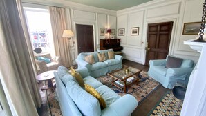 Un salon avec des canapés bleu clair, une table basse et un tapis à motifs. On aperçoit un piano et une porte au fond.