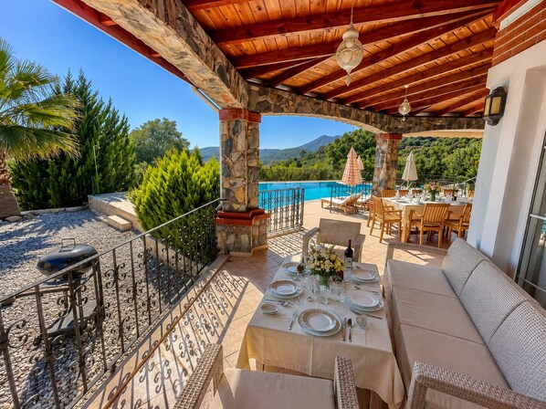 An outdoor dining area with a pool view, stone pillars, and a wooden ceiling.