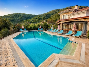 A rectangular pool with blue tiles, surrounded by lounge chairs and umbrellas, with a stone pathway leading to a building with a stone facade and a balcony.