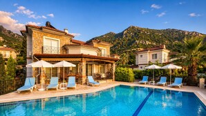 A poolside area with lounge chairs and umbrellas, a stone building with a tiled roof, and a mountainous backdrop. - Stunning Villa Sana Perfect Condition, Private 100 m³ Pool & 1000m2 Garden WIFI (Muğla)