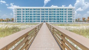A modern building with a long wooden boardwalk leading to it, surrounded by sand and sparse vegetation.