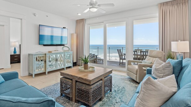 A spacious living room with a large TV, comfortable seating, a ceiling fan, and a view of the ocean through large windows.