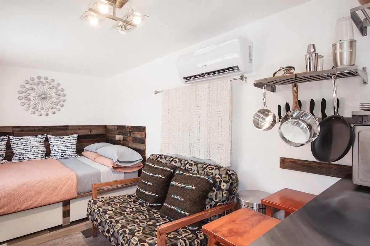 A compact room with a bed, a patterned sofa, a small table, and a kitchenette area with pots and pans hanging above.
