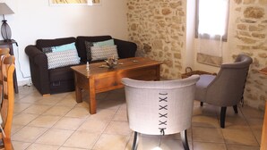 A living room with a stone wall, a wooden coffee table, a grey armchair, and a black sofa with patterned cushions. - SPACIOUS GITE FOR 4 PEOPLE IN AUTHENTIC PROVENCAL FARMHOUSE (Suzette)