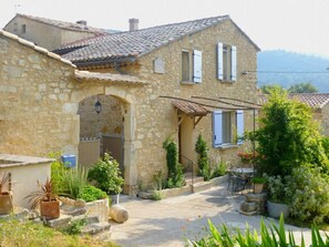A stone house with a tiled roof, a covered porch, and a garden with potted plants and a table.