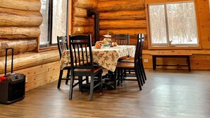 Interior de una cabaña de troncos con comedor, paredes de madera, ventanas amplias y un candelabro.