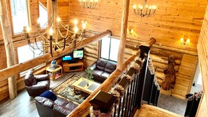 Interior de una cabaña de troncos con techo de madera, candelabro, sofás de cuero, televisor y chimenea.