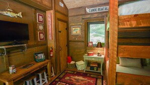 Interior de una cabaña con televisión, paredes de madera, letrero de alquiler de canoa, chimenea y ventana con vista a árboles.