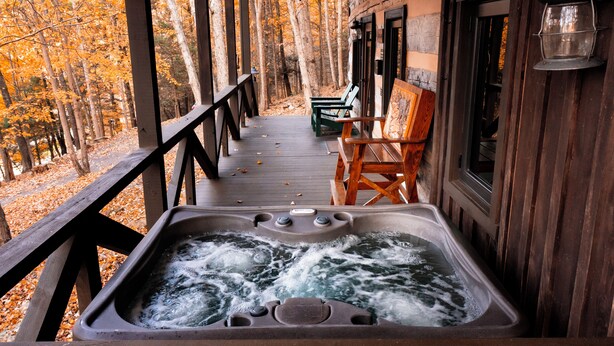 A wooden deck with a hot tub, wooden chairs, and a view of a forest. - The Wilderness Lodge at Four Fillies Lodge w/ Hot Tub (Peterstown)