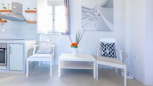 A modern, minimalist living room with white furniture, a kitchen area, and a large window with curtains. - Villa Olena: 2 villas 10 pax 2 pools Punda Beach Paros (Paros)