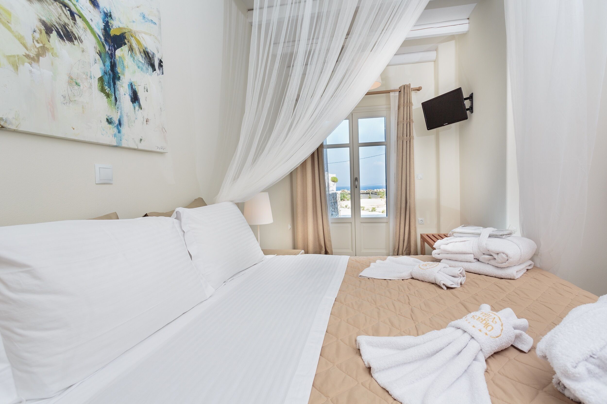 A hotel room with a large bed, a TV mounted on the wall, and a view of the sea through a door.