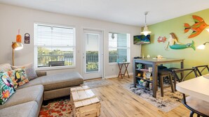 Una sala de estar acogedora con un sofá, una mesa de centro y una mesa de comedor. Hay tres ventanas con persianas, un ventilador de techo y una pared con un mural de peces.