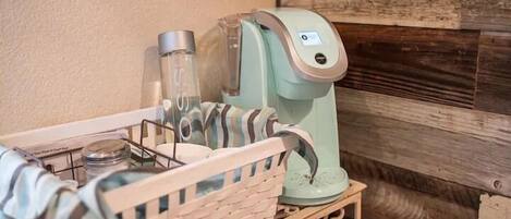Una cafetera sobre un estante de madera con una cesta que contiene un paño y un recipiente de vidrio.