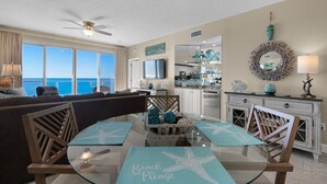 A spacious living area with a large window offering a view of the ocean, a dining table with starfish placemats, and a kitchen with modern appliances.