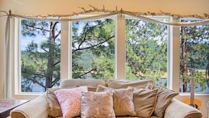 A beige sofa with patterned cushions in front of a window with a view of pine trees.