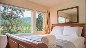 A bedroom with a wooden bed, a large window with a view of trees and hills, a bedside lamp, and a mirror on the wall.