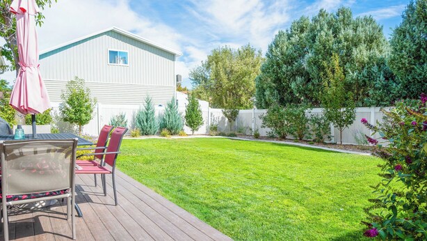 A backyard with a wooden deck, a white building, and a variety of trees and shrubs.