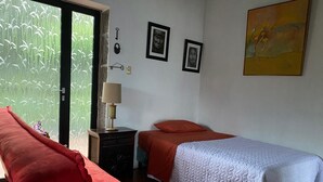 Ein Zimmer mit einem roten Sofa, einem Bett mit orangefarbener Decke, einem weißen Bettüberwurf, einem Nachttisch mit Lampe und einem Fenster mit Bambusmuster.