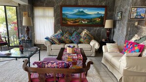 Ein Wohnzimmer mit einem hölzernen Couchtisch, der mit bunten Textilien bedeckt ist, einer Couch mit gemusterten Kissen und einem Bild an der Wand.